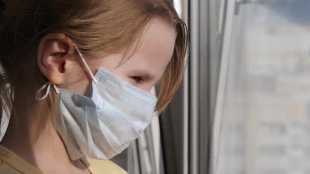 Karantän, hot om coronavirus. Sorgligt barn i skyddande medicinsk mask sitter på fönsterbrädan och tittar ut genom fönstret. Virusskydd, pandemi, förebyggande epidemi. — Stockvideo