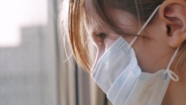 Karantän, hot om coronavirus. Sorgligt barn i skyddande medicinsk mask sitter på fönsterbrädan och tittar ut genom fönstret. Virusskydd, pandemi, förebyggande epidemi. — Stockvideo