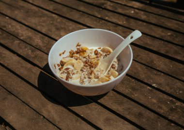 Tahta bir bankta kaşıkla beyaz kasede muzlu müsli.