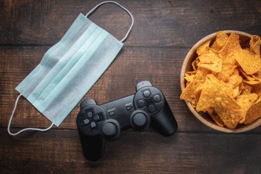 Oyun konsolu RC, bir kase cips ve ahşap masada tıbbi maske. Coronavirus Covid-19 karantinaya alınmıştır..