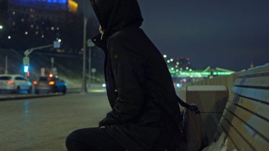 Kapüşonlu adam bankta sırasında karlı winte gece sokakta oturuyor