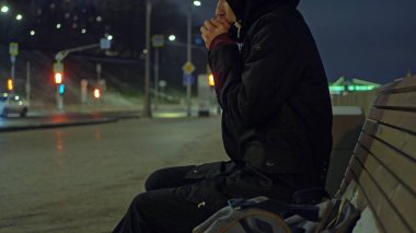 Kapüşonlu adam bankta gece sokakta sırasında karlı winte ısıtır