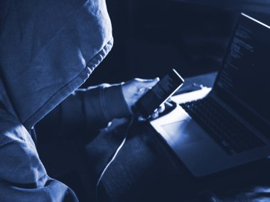 Kaputta oturan isimsiz kişi bilgisayarın önünde oturuyor ve cep telefonu ve dizüstü bilgisayarla çalışıyor.