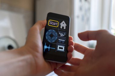 İnsan eli akıllı telefonu tutuyor ve mutfaktaki akıllı ev uygulamasını kullanıyor.