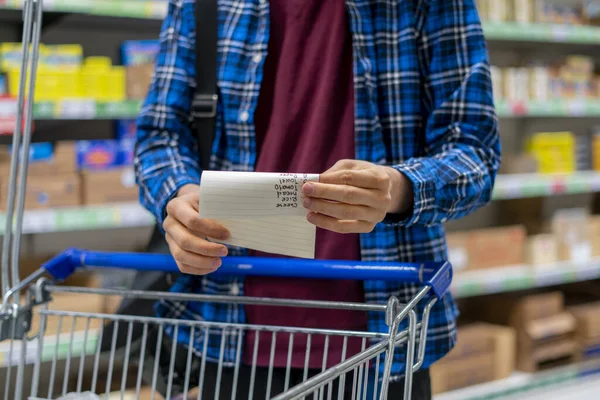 A person's hands holding a shopping list paper sheet and check buying products in grocery store — ストック写真