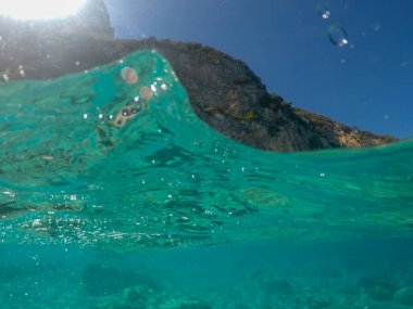 Su yüzeyinin üstünde ve altında, kayalar ve mavi gökyüzü, Akdeniz, Cala dei Gabbiani, Sardinya, İtalya 'daki su hattı ile bölünmüş