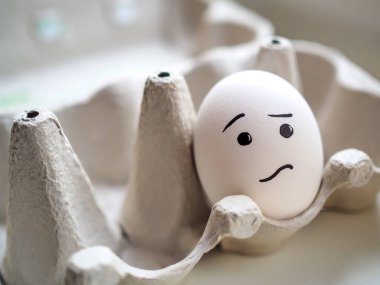 Lonely white egg with sad emoji lies in egg box clipart