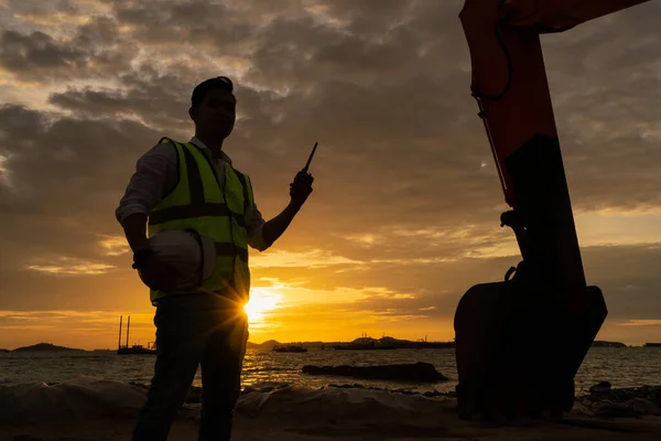 Construction engineer holding radio communications with backhoe in construction site by the sea at sunset. Engineer Concept.