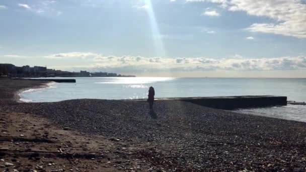 Девочка в красном пальто бегает по морю, наслаждаясь солнечными лучами — стоковое видео