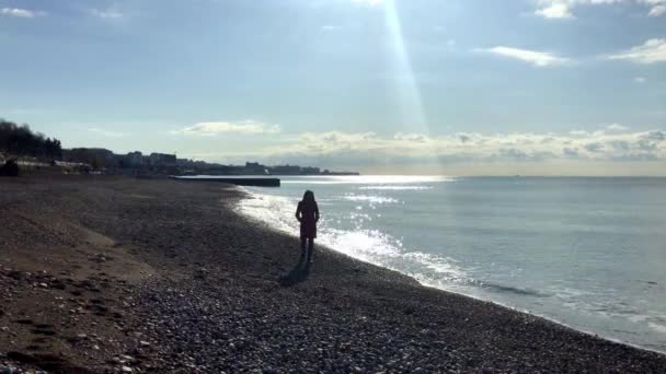Dziewczyna w czerwonym płaszczu biegająca po morzu i ciesząca się promieniami słońca — Wideo stockowe