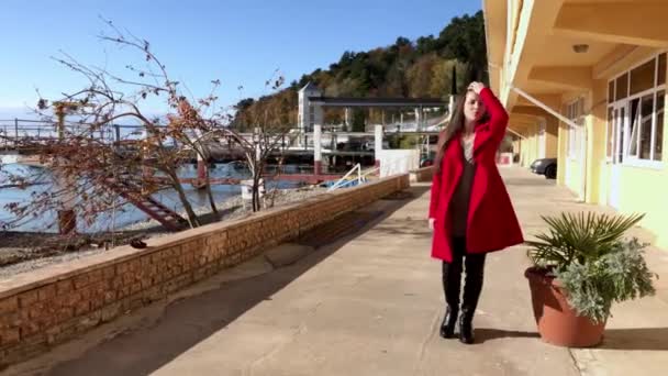 A piros kabátos lány tiszta napos időben sétál a parton. — Stock videók