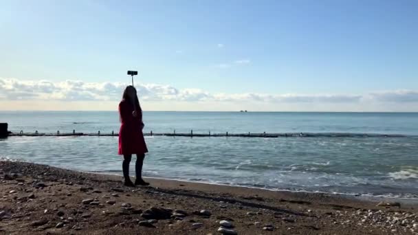 Uma mulher com um pau de selfie caminha à beira-mar, grava um vídeo para o blog — Vídeo de Stock
