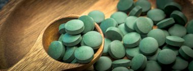 Tabletlerdeki yeşil yosunlar - klorella, tahta zemin üzerinde tahta kaşıkta spirulina - yakın plan pankart tasarımı.