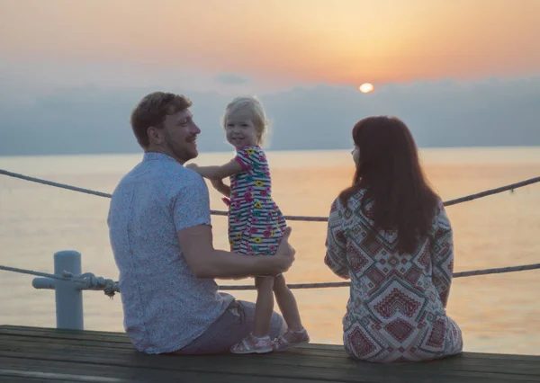 Mom, Dad and daughter are watching the dawn on the pier. A girl looks at the camera, parents look at her