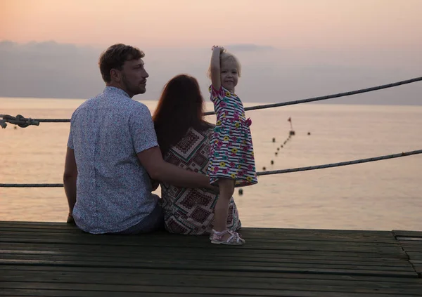 Mom, Dad and daughter are watching the dawn on the pier. The girl looks at the camera, dad looks at her