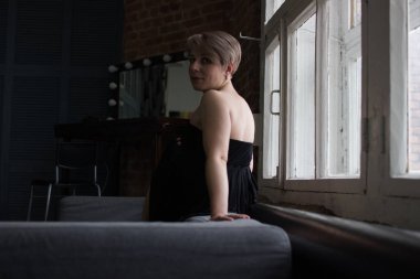 Siyah elbiseli bir kız pencerenin yakınındaki kanepede oturuyor. Sarı saç, kısa saç kesimi, kameraya benziyor