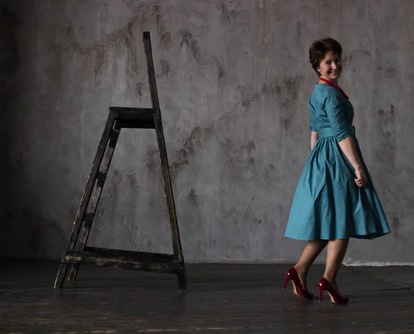 A woman in a turquoise dress in red shoes stands dancing next to the stairs on a light gray background