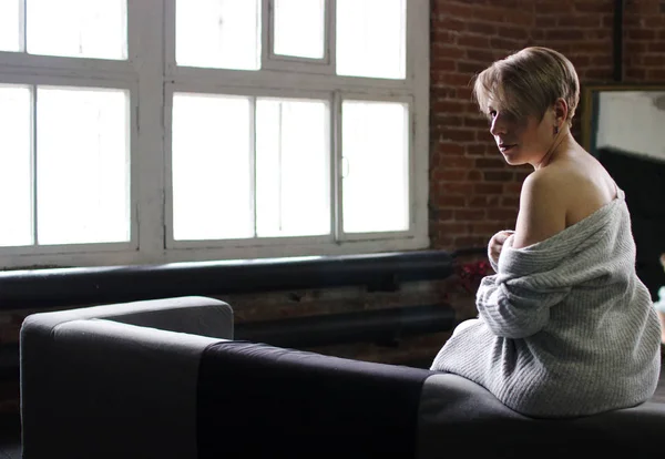 Young Woman Sits Gray Sofa Bare Shoulders Gray Sweater Brick — Stock Photo, Image