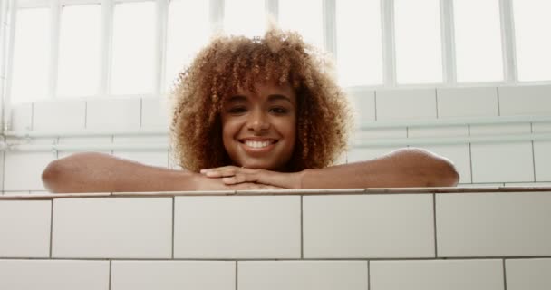 Porträt einer schwarzen Frau mit lockigem Haar, die sich an Bord eines Swimmingpools lehnt und in die Kamera schaut — Stockvideo