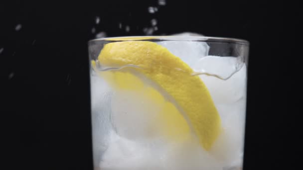 Glass with ice and lemon slice on black with homemade lemonade pouring in it — Stock Video