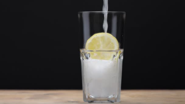 Склянка з льодом і лимонним шматочком на чорному з домашнім лимонадом, що вливається в нього — стокове відео