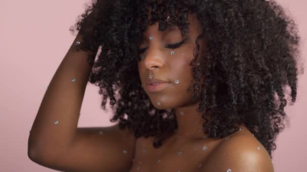 Mujer negra de raza mixta con pelo rizado cubierto de maquillaje de cristal sobre fondo rosa en el estudio — Vídeos de Stock