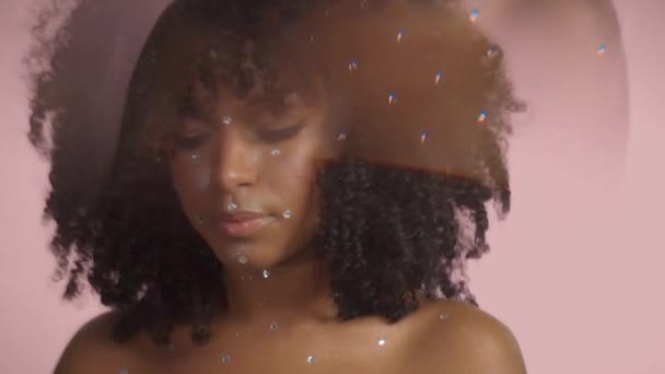 Mujer negra de raza mixta con pelo rizado cubierto de maquillaje de cristal sobre fondo rosa en el estudio — Vídeos de Stock