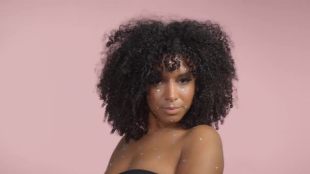 Mulher preta de raça mista com cabelo encaracolado coberto por maquiagem de cristal em fundo rosa em estúdio — Vídeo de Stock