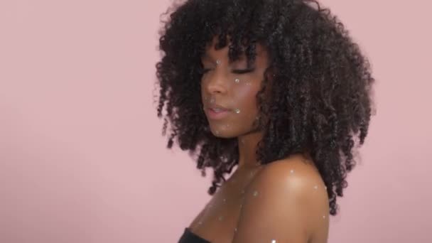 Черная женщина смешанной расы с вьющимися волосами, покрытая хрустальным макияжем на розовом фоне в студии — стоковое видео