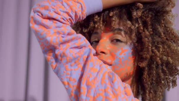 Gemengde race vrouw met krullend haar en heldere neon make-up patroon op het gezicht hetzelfde als op haar sweater — Stockvideo