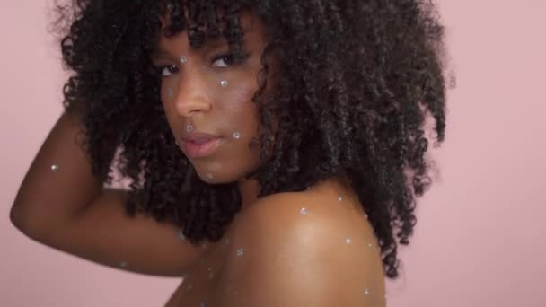 Mieszane rasy czarna kobieta z kręconymi włosami pokryte kryształowym makijażem na różowym tle w studio — Wideo stockowe