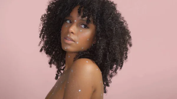 Gemengd ras zwarte vrouw met krullend haar bedekt met kristal make-up op roze achtergrond in de studio — Stockfoto