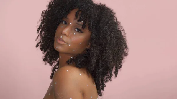 Mixed race black woman with curly hair covered by crystal makeup on pink background in studio — Stock Photo, Image