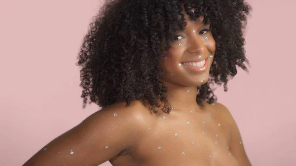 Blandad ras svart kvinna med lockigt hår täckt av kristall makeup på rosa bakgrund i studio — Stockfoto