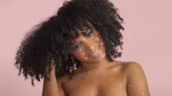 Mujer negra de raza mixta con pelo rizado cubierto de maquillaje de cristal sobre fondo rosa en el estudio — Foto de Stock