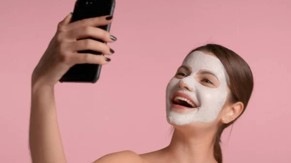 Mujer morena de 30 años con una máscara de arcilla facial en la fabricación de selfie con un teléfono inteligente — Foto de Stock