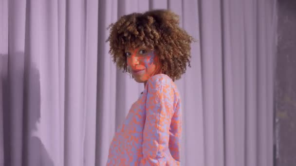 Mulher de raça mista com cabelo encaracolado e padrão de maquiagem neon brilhante no rosto o mesmo que em sua camisola — Vídeo de Stock