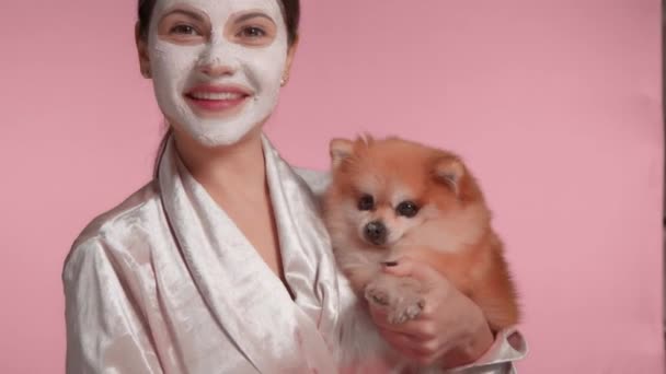 30s donna bruna con argilla maschera facciale sulla tenuta di un piccolo cane soffice in mano — Video Stock