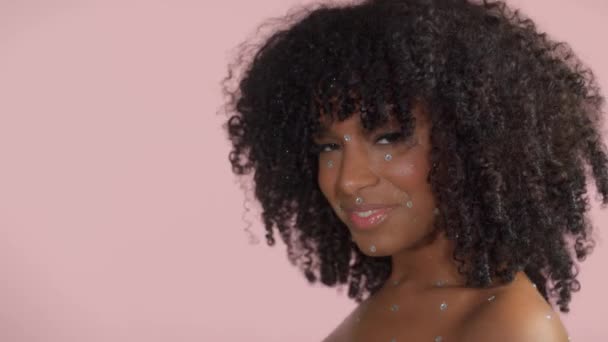 Mixed race black woman with curly hair covered by crystal makeup on pink background in studio — Stock Video