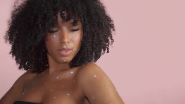 Mujer negra de raza mixta con pelo rizado cubierto de maquillaje de cristal sobre fondo rosa en el estudio — Vídeos de Stock