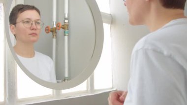 Banyodaki genç kadın aynaya yaklaşır ve yüzüne ve saçlarına dokunur.