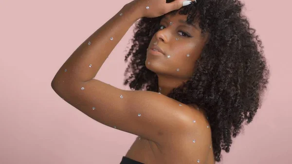 Mujer negra de raza mixta con pelo rizado cubierto de maquillaje de cristal sobre fondo rosa en el estudio — Foto de Stock