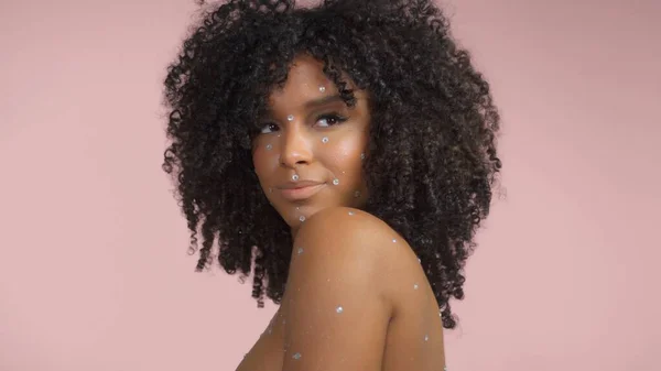 Mujer negra de raza mixta con pelo rizado cubierto de maquillaje de cristal sobre fondo rosa en el estudio — Foto de Stock
