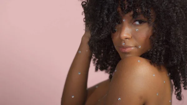 Mujer negra de raza mixta con pelo rizado cubierto de maquillaje de cristal sobre fondo rosa en el estudio — Foto de Stock