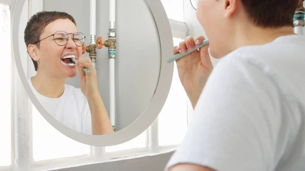 Mujer caucásica con un corte de pelo corto delante del espejo en el baño lleno de luz solar cepillarse los dientes — Foto de Stock