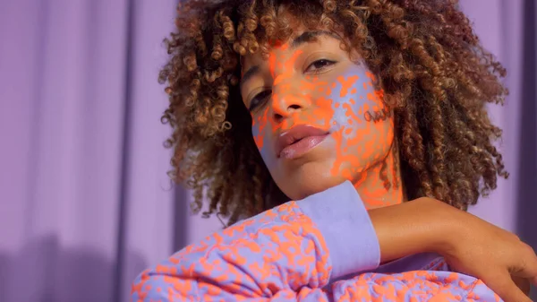 Gemengde race vrouw met krullend haar en heldere neon make-up patroon op het gezicht hetzelfde als op haar sweater — Stockfoto