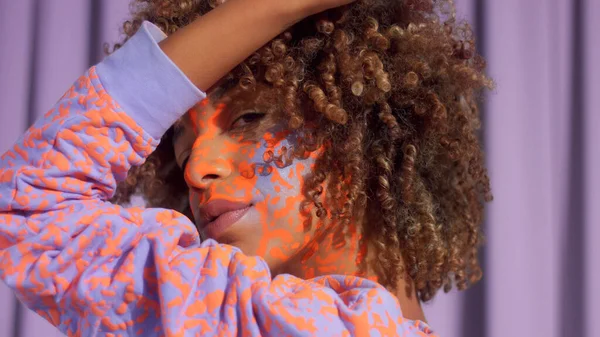 Gemengde race vrouw met krullend haar en heldere neon make-up patroon op het gezicht hetzelfde als op haar sweater — Stockfoto