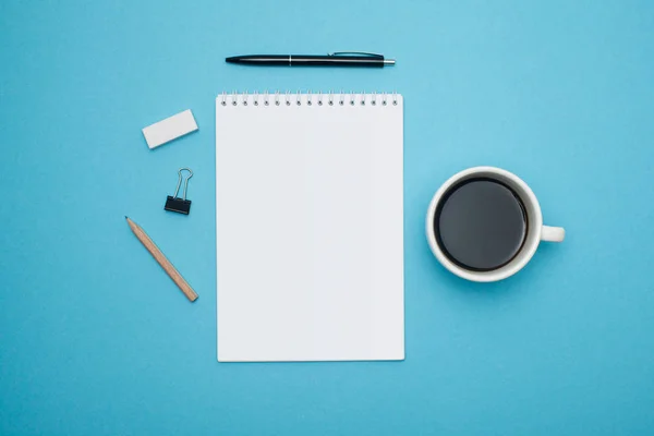 Plat conception laïque de bureau avec des lunettes de crayon cahier blanc café sur fond bleu — Photo