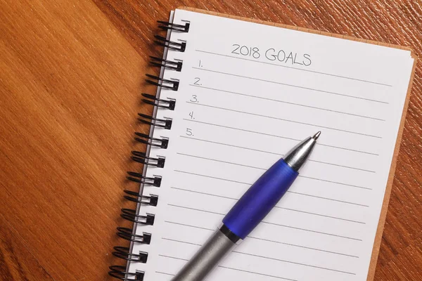 Top view or flatlay of notebook with words 2018 GOALS and blank checklist on wooden table with pen