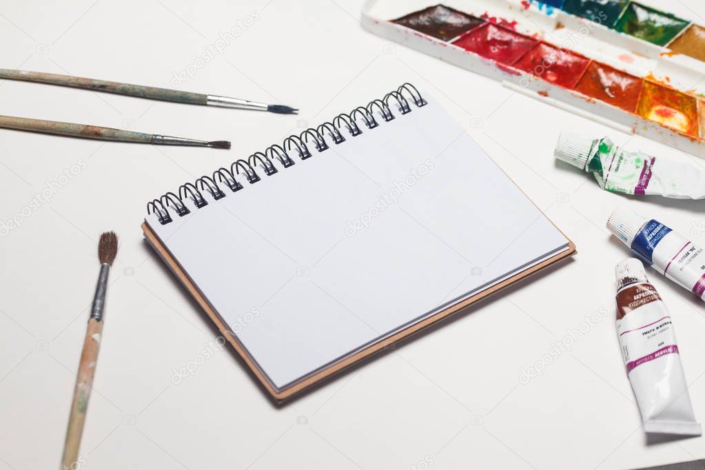 Notebook mock up for artwork with watercolor paints, palette and a paintbrushes. Top view. Artistic work tools on white background.